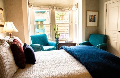 Guest room with two large bright windows, two teal sitting chairs and luxurious bed