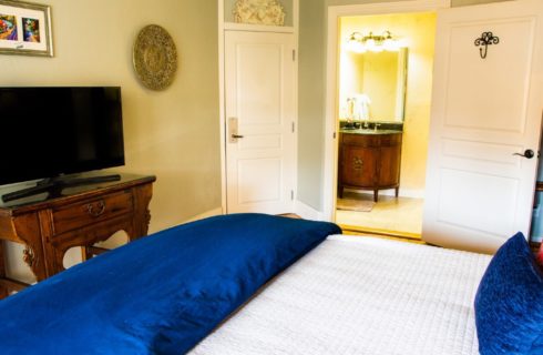 Guest room showing bed with white and blue bedding, TV console, and doorway into bathroom