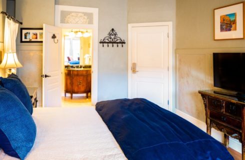 Bed with white and blue bedding in guest room with TV console and doorway into elegant bathroom