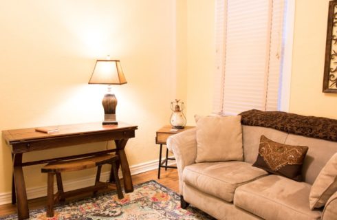 Sitting area with tan and brown couch, writing desk with lamp and decorative art on the wall