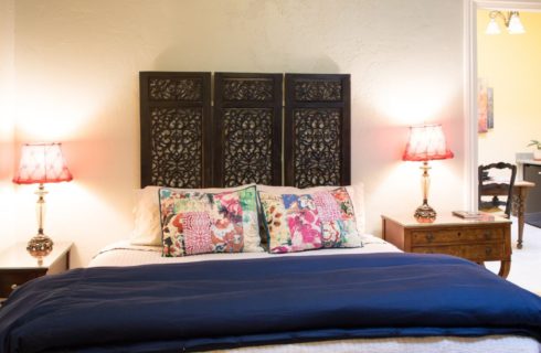 Single bed with white and blue bedding, tall decorative headboard with two side tables and doorway into kitchenette