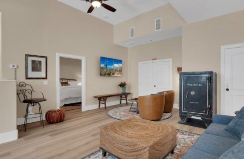 living room with hardwood floors and throw rugs, leather chairs facing flat screened tv on wall, tall black safe, view into bedroom through open door