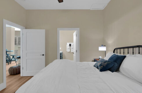 bedroom with hardwood floors and king bed with white comforter, view into bathroom and kitchenette through open doors