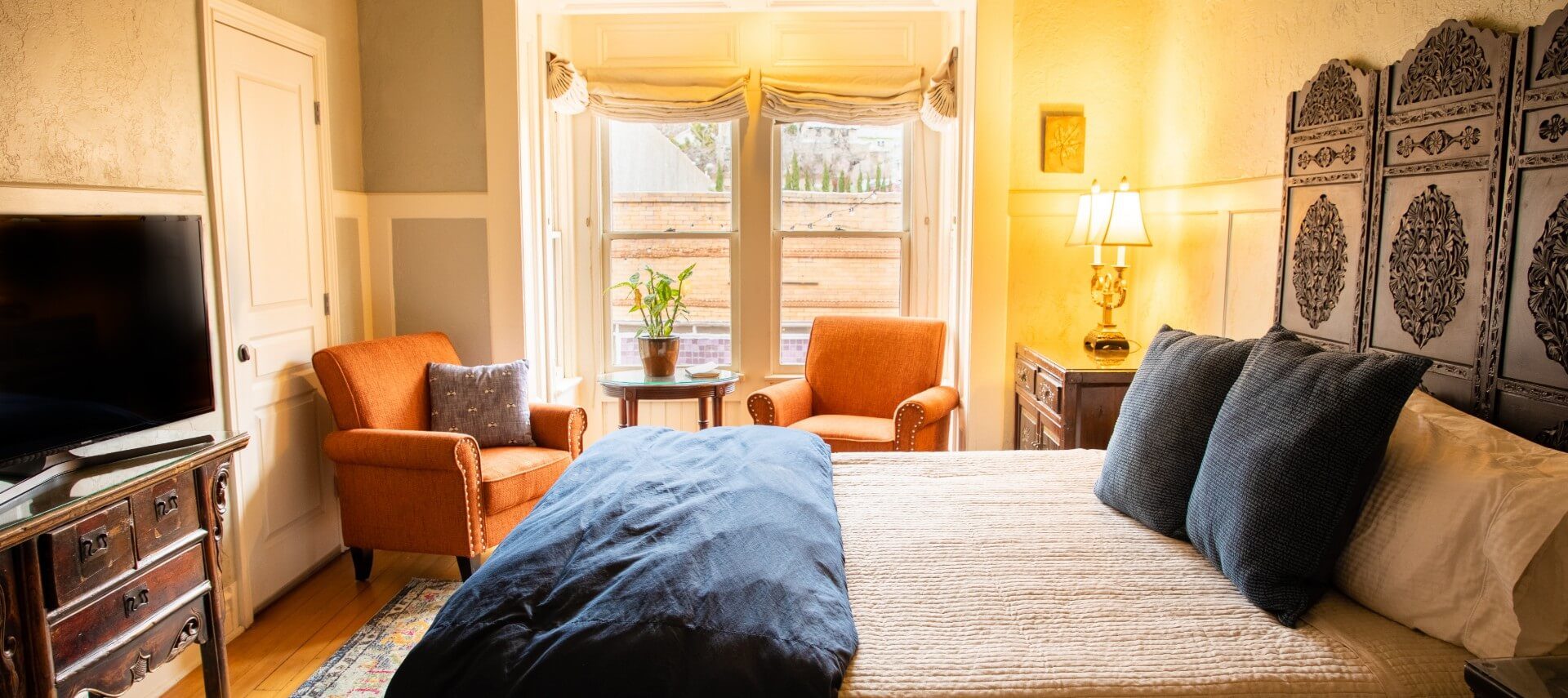 Beautiful room with king bed with decorative headboard, two orang sitting chairs in front of bright windows