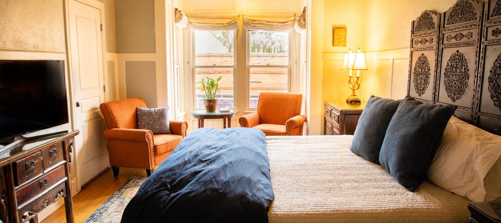 Large bed with decorative headboard in spacious suite with orange sitting chairs and two bright windows