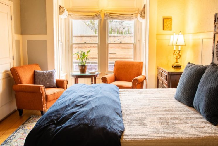 Large bed with decorative headboard in spacious suite with orange sitting chairs and two bright windows