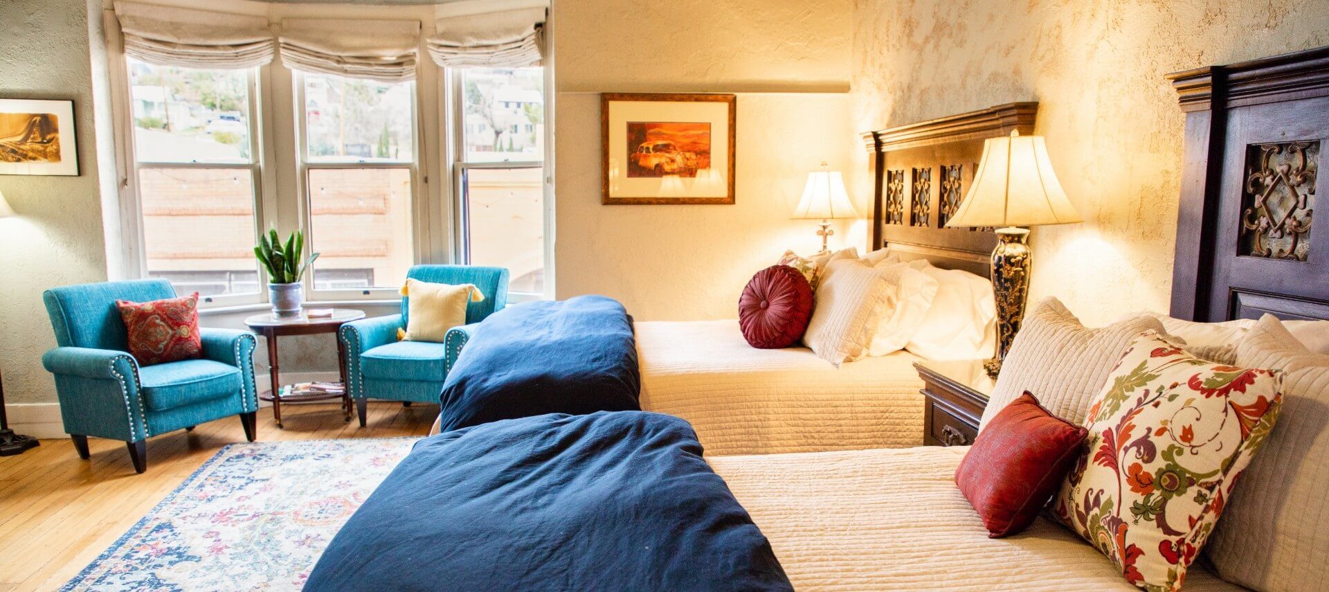 Two queen beds in large guest room with two teal sitting chairs in front of three large windows