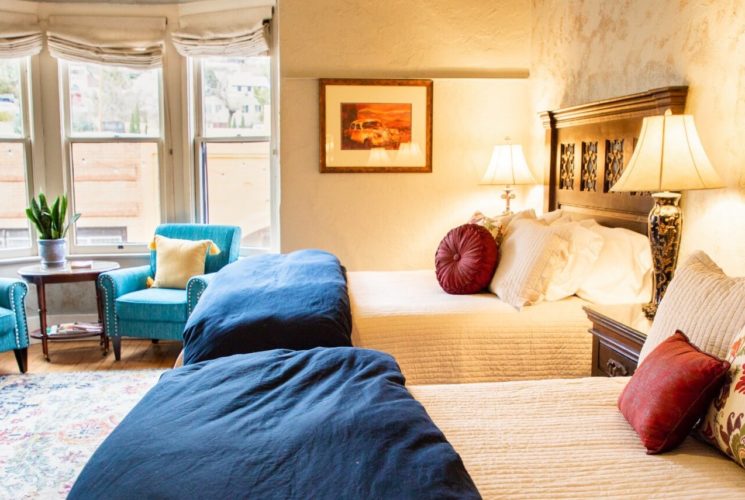 Two queen beds in large guest room with two teal sitting chairs in front of three large windows