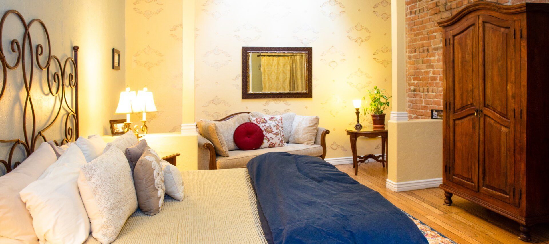 King bed with iron headboard in large room with couch and tall brown armoire against a brick wall