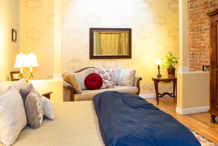 King bed with iron headboard in large room with couch and tall brown armoire against a brick wall