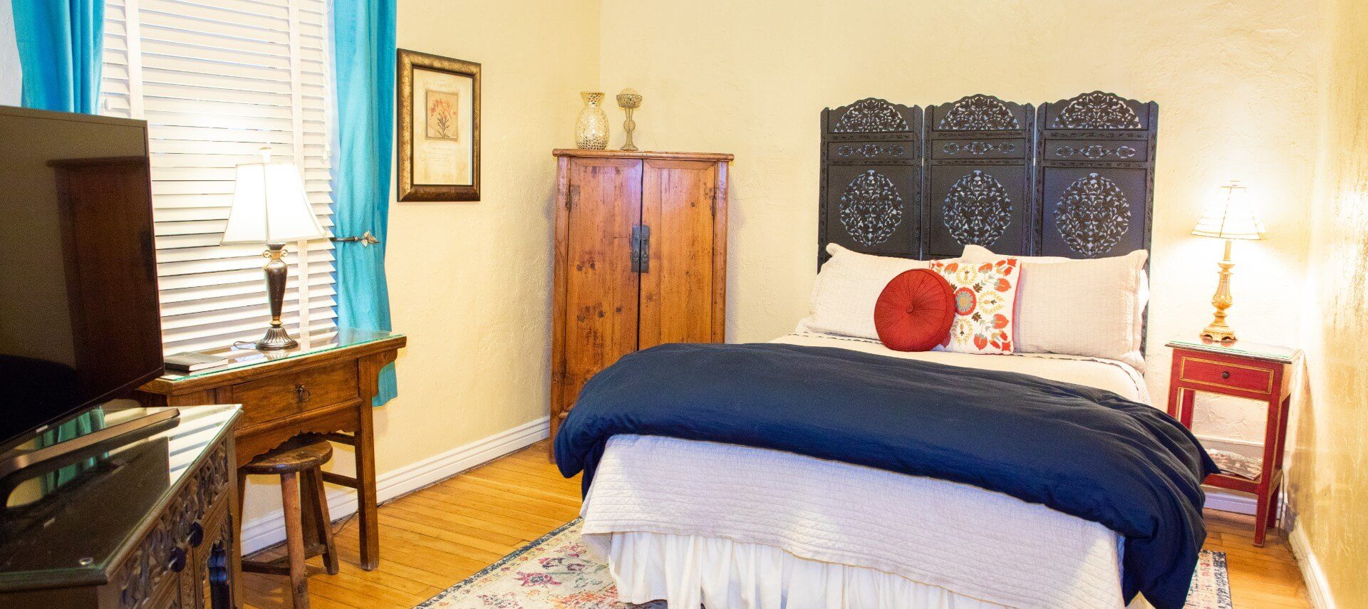 Queen bed with decorative headboard, corner armoire and desk in front of one window