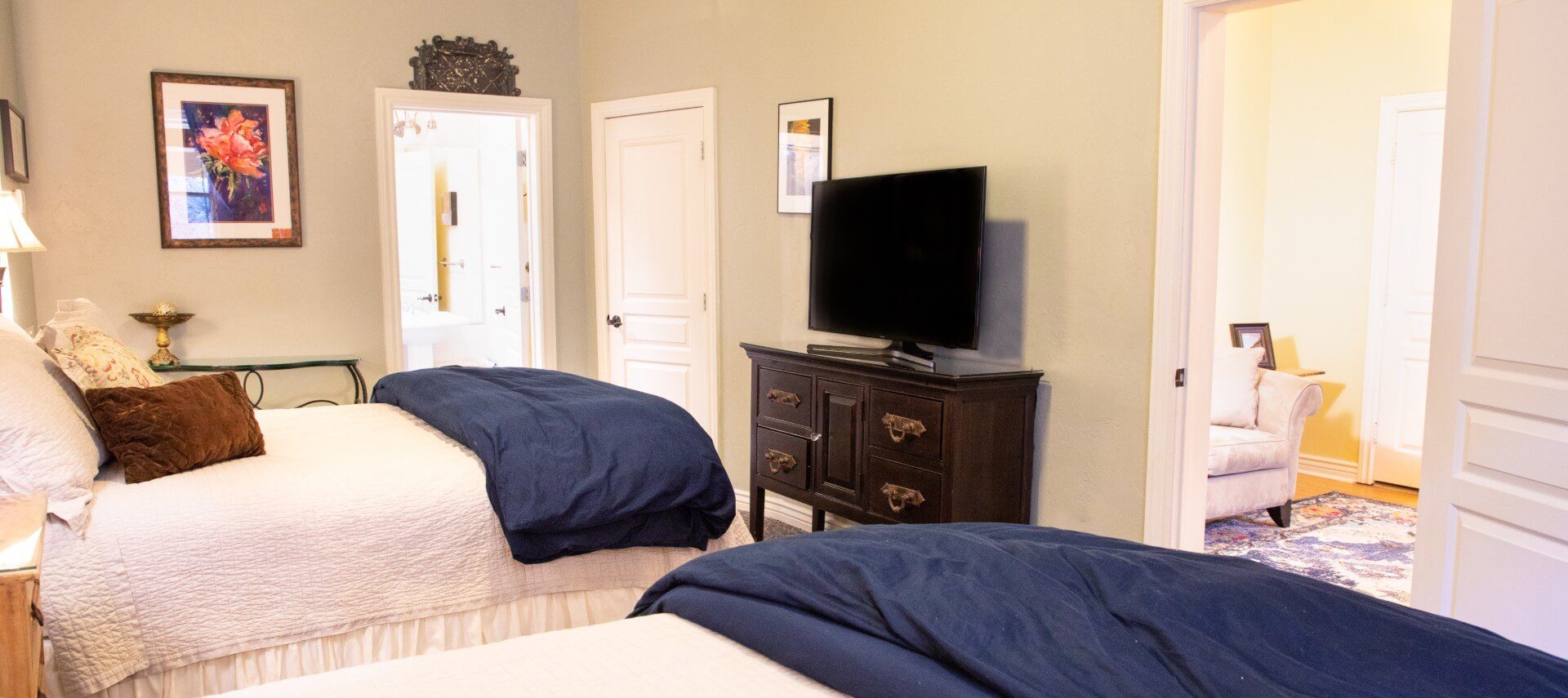 Two queen beds with white and blue bedding in room with TV console and doorway into separate sitting room with couch