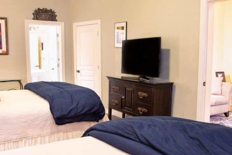 Two queen beds with white and blue bedding in room with TV console and doorway into separate sitting room with couch