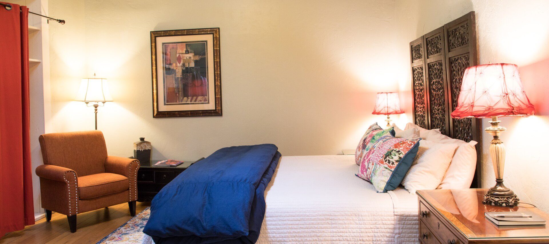 Queen bed and white and blue bedding decorative headboard, sitting chair, and closet with red curtain