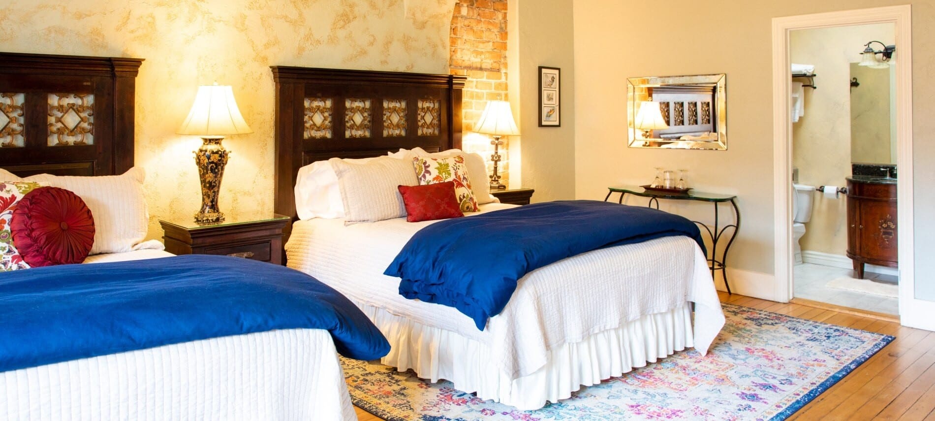 Two queen beds with white and blue bedding in a large guest room with doorway into bathroom