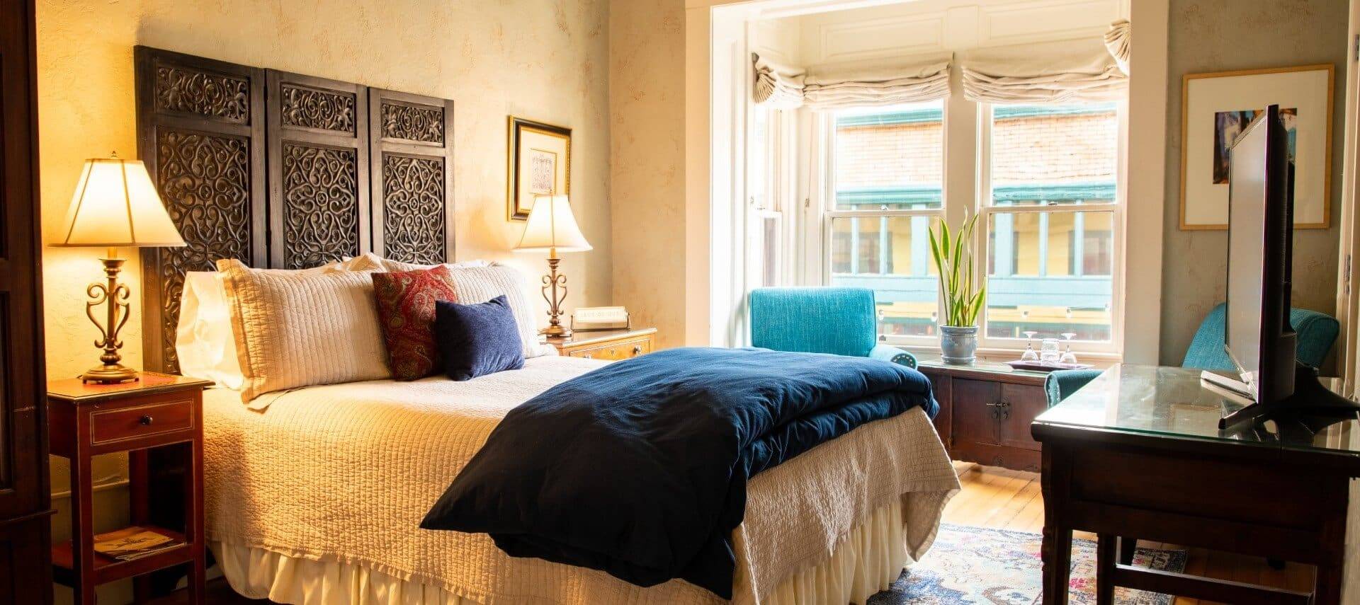 Peaceful room with single bed with blue and white bedding, sittings chairs in front of two large windows and TV console