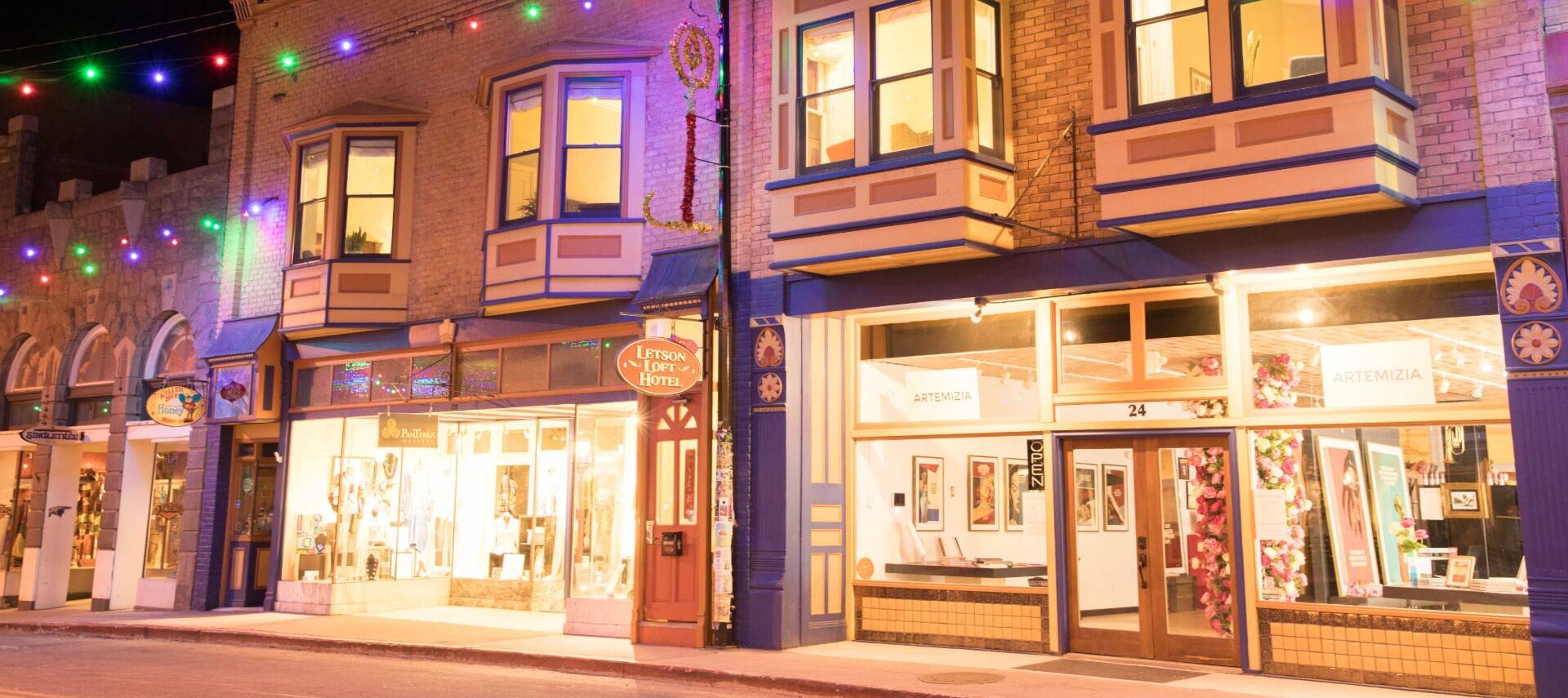 Small town street at night featuring several storefronts with glass windows showing their items for sale
